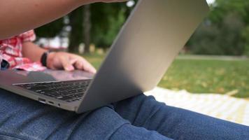 närbild aisan kvinna sitter och använder laptop i parken. öppnar och skriver på laptop, arbetar med bra utsikt över parken. går ut i helgen. gå picknick koncept video