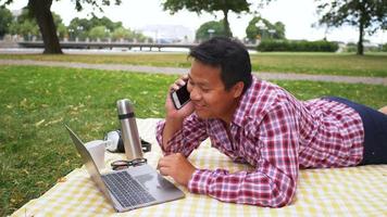 hombre feliz aisan acostado, trabajando en la computadora portátil y teniendo una llamada en el parque. trabajando al aire libre con buena naturaleza, tomando un poco de aire fresco. salir el fin de semana. ir a concepto de picnic video