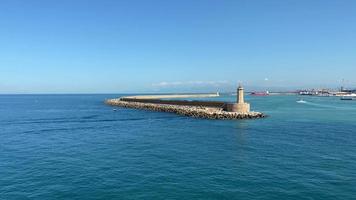 Luchtfoto van een hoge en stevige vuurtoren met rotsen aan de rand van een lange bochtige haven in het midden van de brede blauwe zee tijdens een heldere zonnige dag, langzaam naar rechts draaiend video