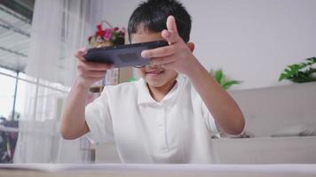 glad liten asiatisk glasögon pojke målar färdig på bok, tar ett foto och skickar till en vän på telefonen i vardagsrummet. avkoppling och hobby koncept. video