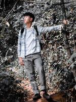Young asian man with a backpack stands photographed by old majestic tree in forest during break from hiking traveling in vacation. Healthy lifestyle concept.