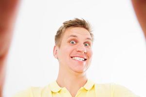 Comic loco en camiseta casual sonriendo a la cámara con el pulgar hacia arriba mientras toma selfie aislado sobre fondo blanco. foto
