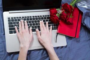 Fashion blogger working with laptop in bed, flat lay, top view. Top view mockup image of a woman sitting on a bed , using and typing on laptop with red notebook and flowers photo