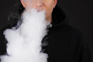 Vaping white man holding a mod. A cloud of vapor. Black background. Vaping an electronic cigarette with a lot of smoke. Vape concept copy space Selective focus photo
