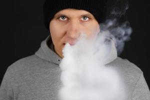 el hombre fuma un cigarrillo electrónico en el fondo oscuro, los hombres con capucha vapean y liberan una nube de vapor. chico con vaping sobre fondo negro foto