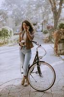 Young woman with mobile phone drink coffee to go by the bicycle on autumn day photo