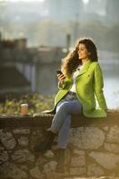 Pretty young woman using smartphone while sitting by the river and drinking takeaway coffee photo