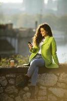 Pretty young woman using smartphone while sitting by the river and drinking takeaway coffee photo