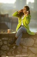 Pretty young woman listening music with smartphone and takeaway coffee by the river on a sunny autumn day photo