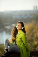 Pretty young woman listening music with smartphone and takeaway coffee by the river on a sunny autumn day photo