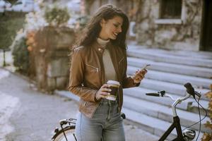 Young woman with mobile phone drink coffee to go by the bicycle on autumn day photo