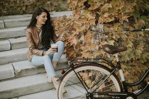 Young woman with mobile phone drink coffee to go at the stairs by the bicycle on autumn day photo