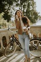 Young woman with mobile phone drink takeaway coffee by the bicycle on autumn day photo