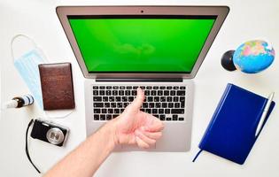 Desktop green screen computer with blue notebook, globe stand, passport, compact camera and hand show thumbs up on white background. Travel international reopening concept. photo