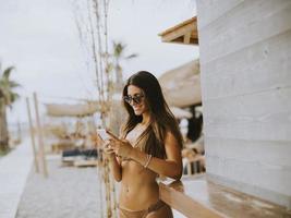 Young woman in bikini standing by the surf cabin on a beach and using mobile phonr at summer day photo