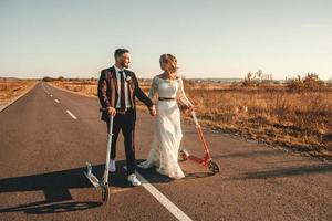 Smiling wedding couple riding a on scooters along the road outside the city at sunset. Place for text or advertising photo
