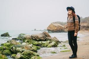 Woman in a hat and scarf with a backpack on coast against background of rocks against beautiful sea