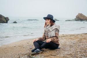 Woman in a hat and scarf sits on coast against background of rocks against beautiful sea photo