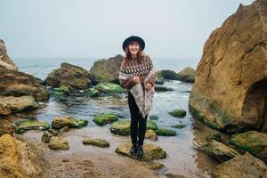 Mujer con sombrero y bufanda de pie sobre una roca frente a un hermoso mar en el horizonte foto