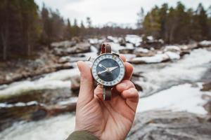 Beautiful male hand holds a magnetic compass against the background of mountain river, rocks and forest. The concept of finding yourself the way and the truth