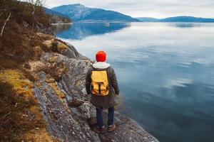 Young man with a yellow backpack wearing a red hat standing on a rock on the background of mountain and lake. Space for your text message or promotional content. Travel lifestyle concept photo