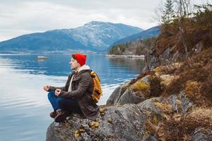 Traveler man in a meditative position sitting on a rocky shore on the background of a mountain and a lake. Space for your text message or promotional content