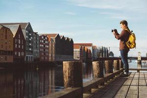 Fotógrafo con mochila amarilla de pie en el muelle de madera tomando fotos contra el fondo de la ciudad y el río