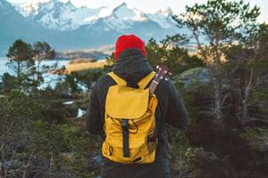 Traveler man is standing in the middle of a forest with a guitar on background of mountains and lake. Wearing a yellow backpack in a red hat. Place for text or advertising. Shoot from the back