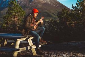 Young man playing guitar sitting on a wooden table against the background of mountains, forests and lakes. Relaxing and enjoying sunny days. Place for text or advertising photo