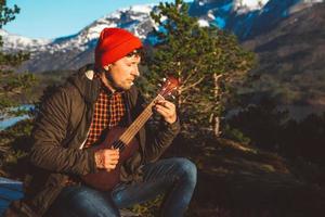Young man playing guitar sitting on a wooden table against the background of mountains, forests and lakes. Relaxing and enjoying sunny days. Place for text or advertising photo