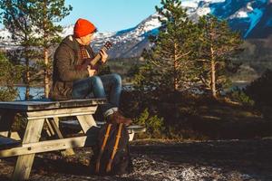 Young man playing guitar sitting on a wooden table against the background of mountains, forests and lakes. Relaxing and enjoying sunny days. Place for text or advertising photo