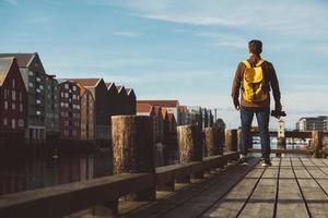 Fotógrafo con mochila amarilla de pie en el muelle de madera tomando fotos contra el fondo de la ciudad y el río