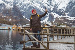 Hombre viajero tomando autorretrato con un teléfono inteligente en el contexto de montañas nevadas, rocas y lagos de pie sobre un muelle de madera. lugar para texto o publicidad foto