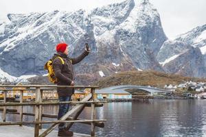 Traveler man taking self-portrait with a smartphone against the background of snowy mountains, rocks and lakes standing on a wooden pier. Place for text or advertising photo