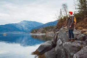 Young man with a yellow backpack wearing a red hat standing on a rock on the background of mountain and lake. Space for your text message or promotional content. Travel lifestyle concept
