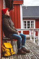 Traveler man with a yellow backpack wearing sits near the wooden red colored house. Travel lifestyle concept.