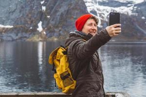 Hombre viajero tomando autorretrato con un teléfono inteligente en el contexto de montañas nevadas, rocas y lagos de pie sobre un muelle de madera. lugar para texto o publicidad foto