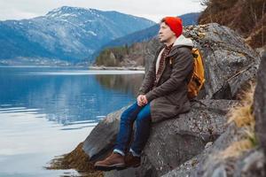 Young man with a yellow backpack wearing a red hat sitting on the shore on the background of mountain and lake. Space for your text message or promotional content. Travel lifestyle concept.