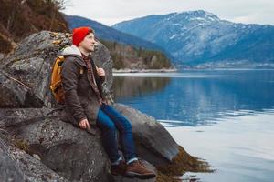 joven con una mochila amarilla con un sombrero rojo sentado en la orilla en el fondo de la montaña y el lago. espacio para su mensaje de texto o contenido promocional. concepto de estilo de vida de viaje. foto