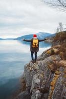 Young man with a yellow backpack wearing a red hat standing on a rock on the background of mountain and lake. Space for your text message or promotional content. Travel lifestyle concept