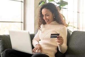 Mujer latina con portátil y mano sujetando la tarjeta de crédito para ir de compras en el sofá