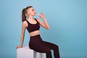 This girl is a fintes coach. She sits on a white cube and drinks water from a bottle. Single tone background photo