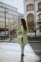 mujer joven usando un teléfono inteligente en la calle y sosteniendo café para llevar foto