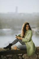 Pretty young woman using smartphone while sitting by the river and drinking takeaway coffee photo
