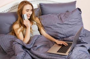 Woman using laptop for store online shopping in bed at home, payment by credit card with online transaction banking. Happy Girl pointed on creditcard.