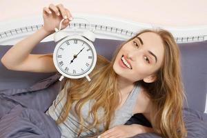 Cute girl holds a clock and lies in bed bedroom. Emotional beautiful woman lying in bed on her day off. photo