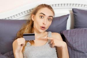 hermosa mujer sosteniendo una tarjeta de crédito y señalando con el dedo. chica sosteniendo una tarjeta bancaria mientras está sentada en la cama en la habitación. foto