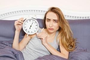 Shocked young woman waking up with alarm, overslept work, bad sleep insomnia. Girl hold clock when lying on bed in the morning photo