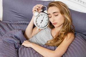 una joven hermosa y somnolienta con los ojos cerrados sostiene un reloj y se acuesta en su cama en el dormitorio por la mañana