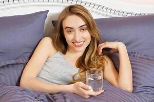 Happy young woman holding a glass of water and lying in bed in the bedroom